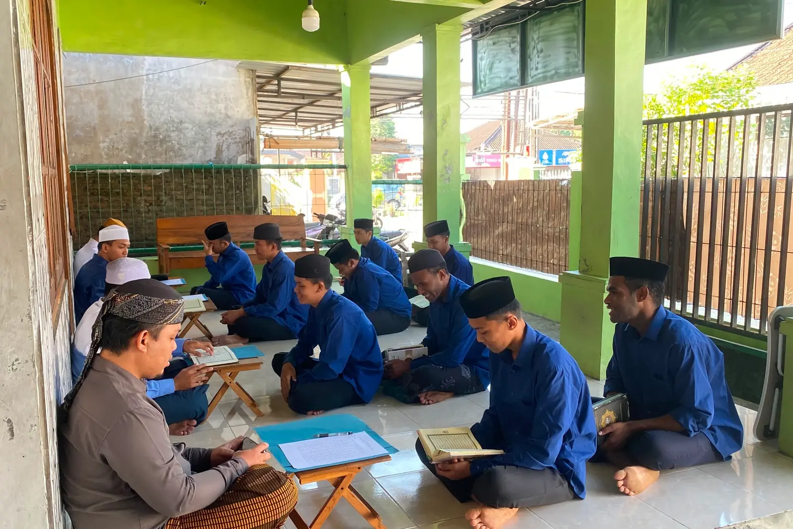 Santri duduk bersila di lantai teras kontrakan dengan meja lipat kecil dan mushaf, seorang ustadz menyimak di sisi kiri