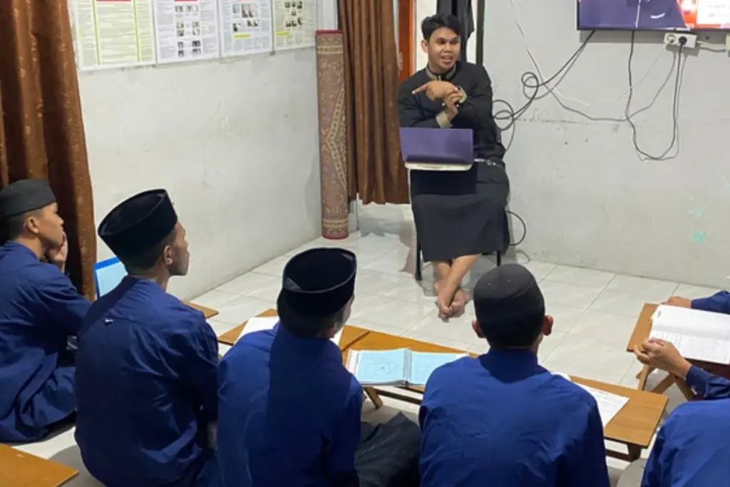 Santri duduk di lantai menghadap pengajar dengan meja lipat kecil, difoto dari belakang barisan santri